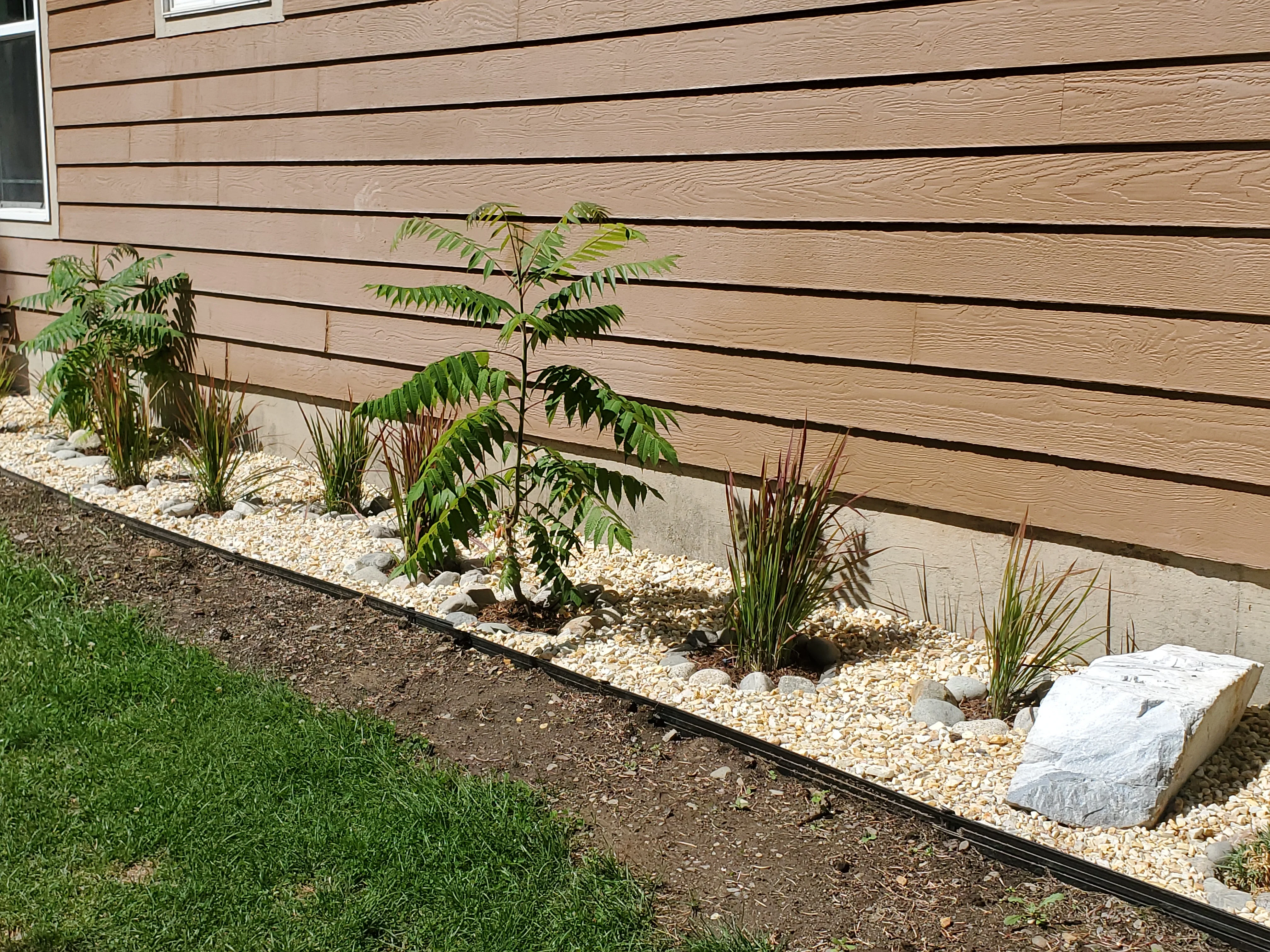 Foundation landscape bed with river rock, ornamental plants, and metal edging along modern home