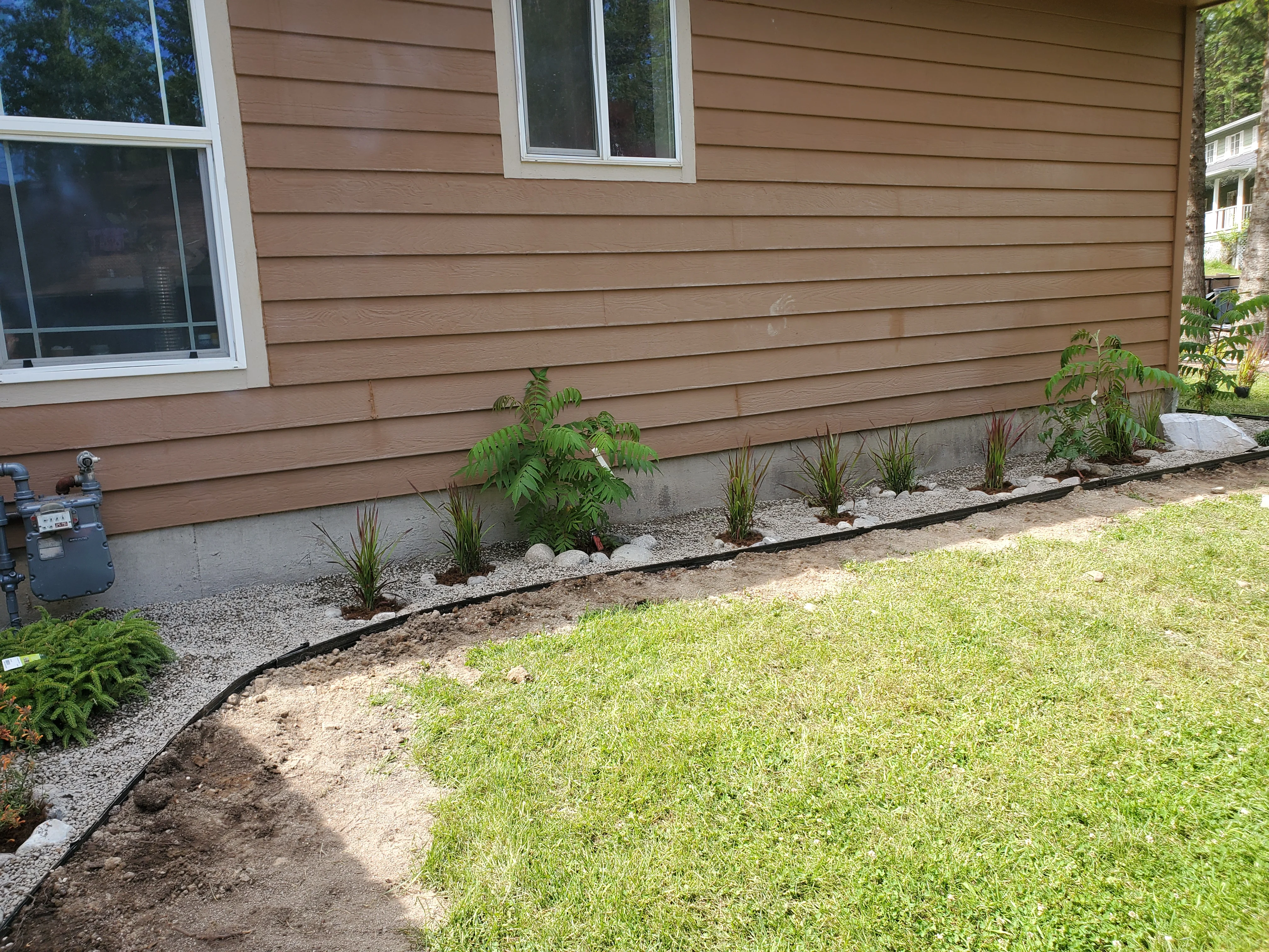 Foundation landscape bed with ornamental grasses, tropical plants, and gravel edging along house