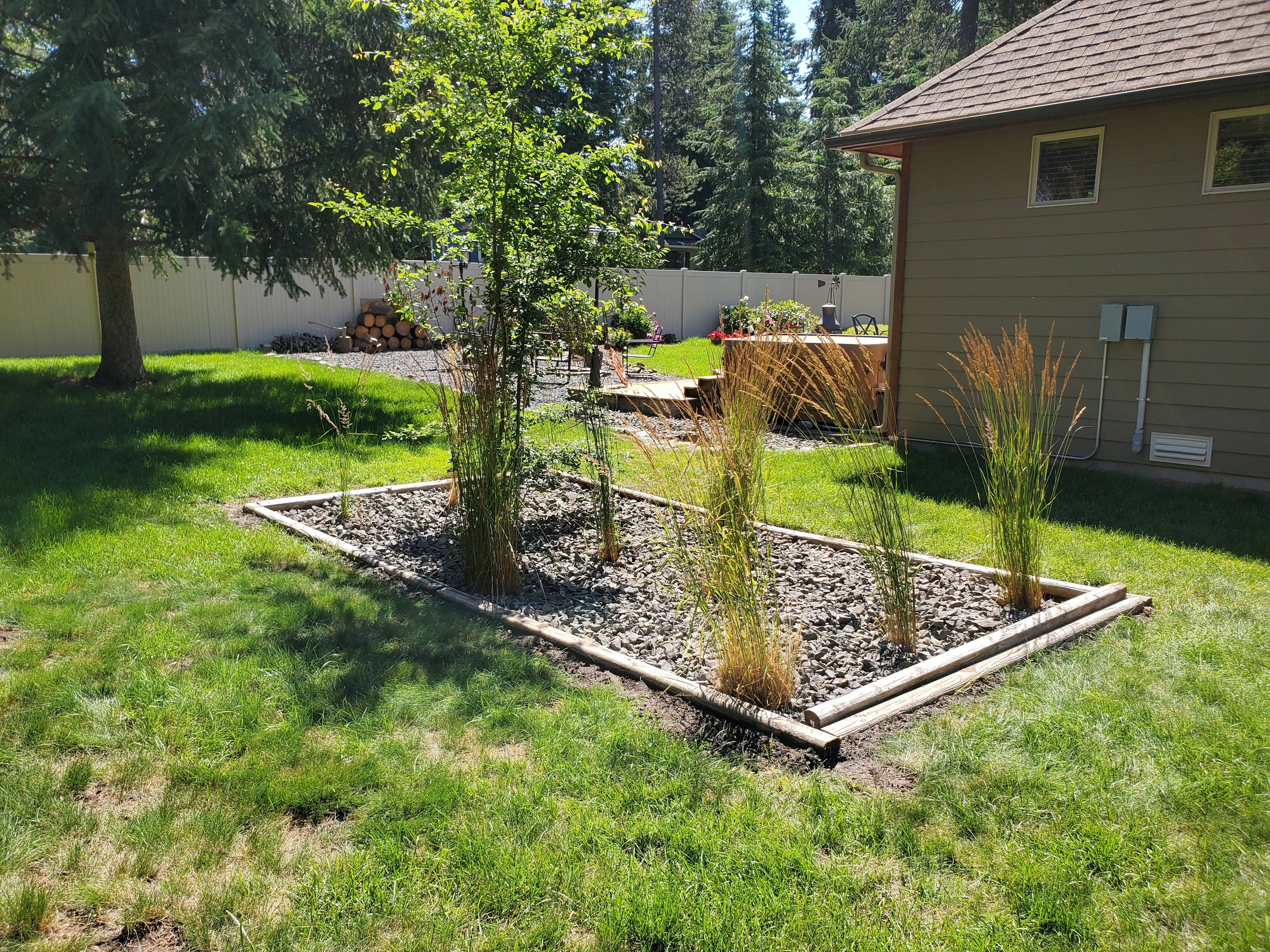 Timber-edged landscape bed with river rock mulch, young tree, and ornamental grasses in residential yard